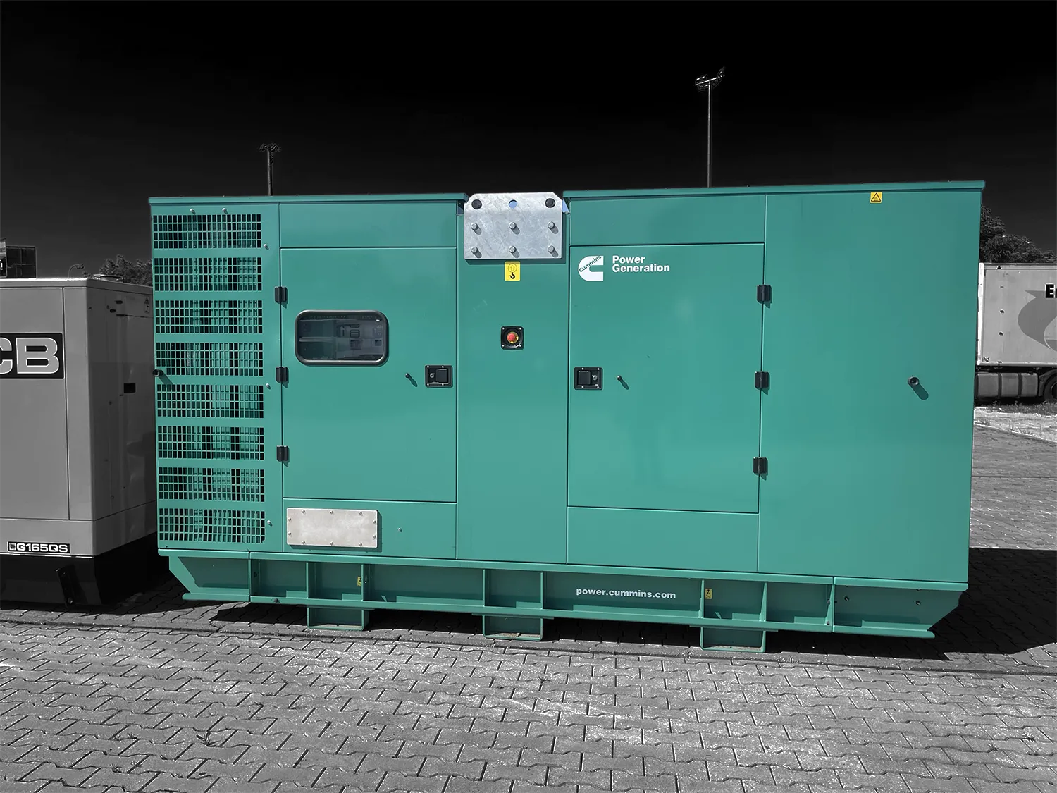Tyrkysový priemyselný dieselový generátor Cummins Power Generation stojí na spevnenej ploche vonku, snímaný zboku na tmavom pozadí.