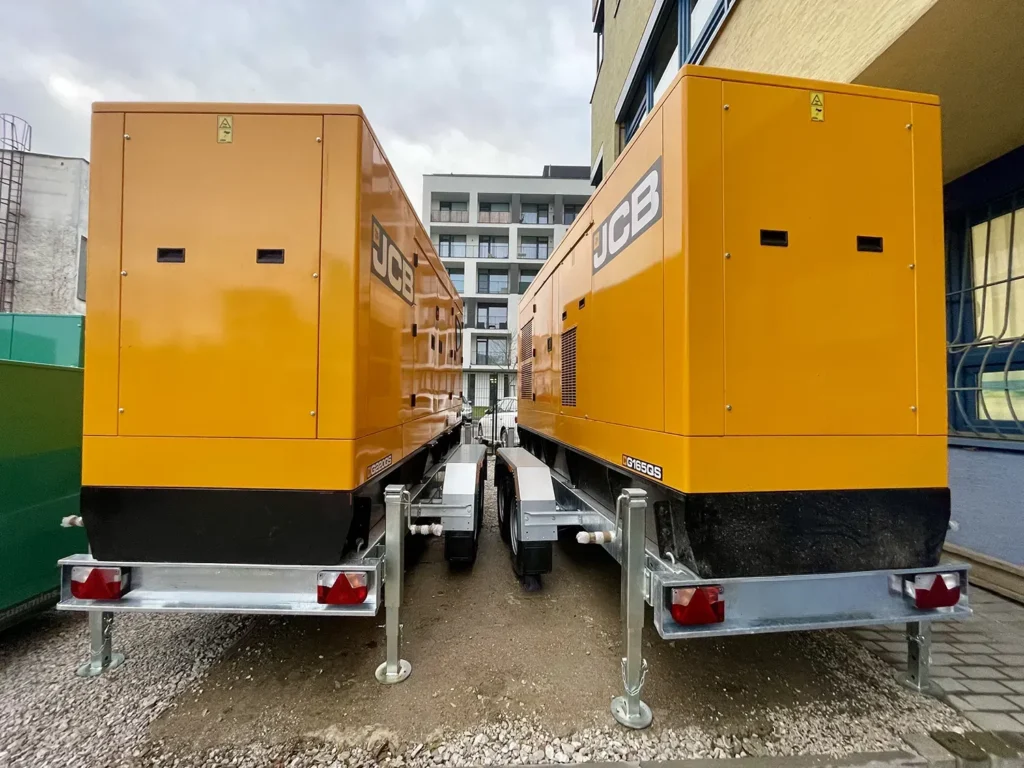 Dva žlté mobilné dieselové generátory JCB na prívesoch stoja vedľa seba vo vonkajšom priestore medzi budovami pod zamračenou oblohou.